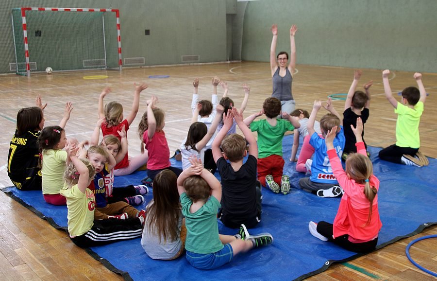 Eine große Kindergruppe sitzt auf Matten und streckt die Arme hoch.