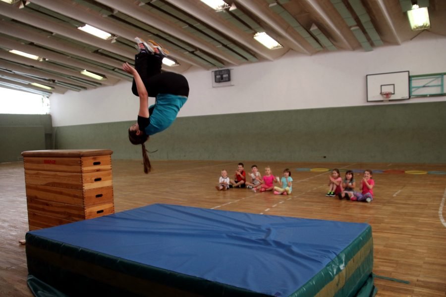 Übungsleiterin Kati Heine springt in der Turnhalle über einen Kasten, dahinter sitzt eine Gruppe Kinder.